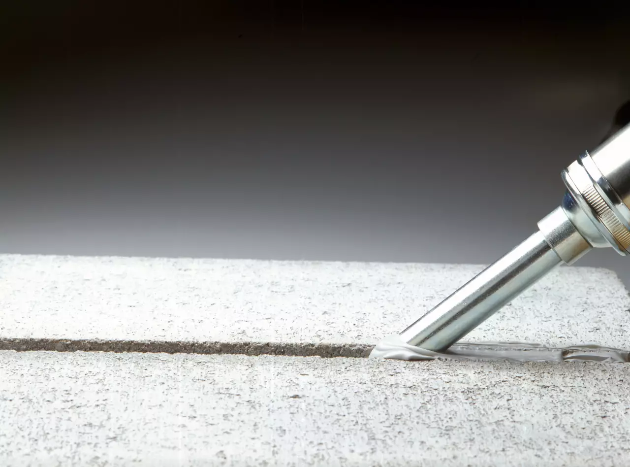 Studio shot of sausage style applicator gun laying bead of Parking Structure Sealant NS (non-slumping) between two pieces of concrete. Product: Sealant NS, Sealant FC. Construction/IBU AProduct brochure