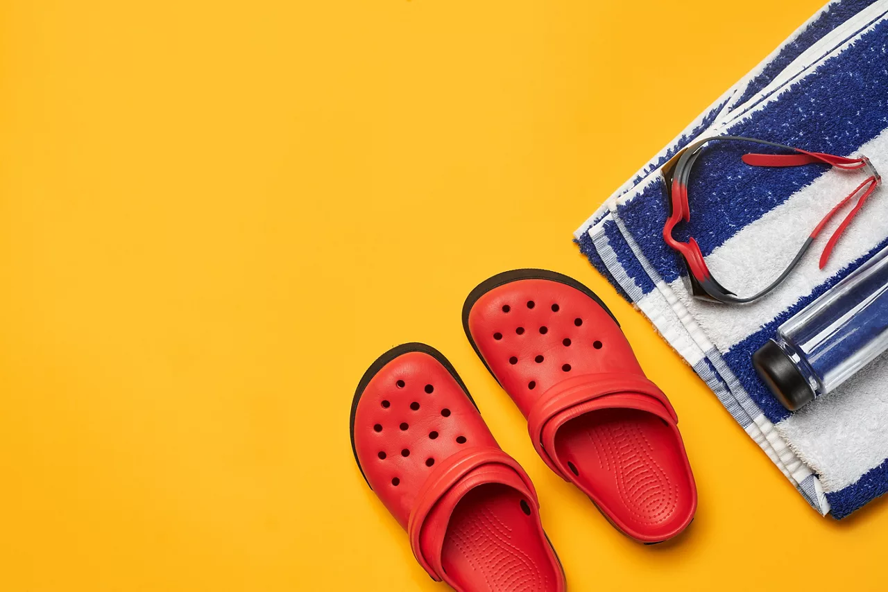 Flat lay, pool accessories such as swimming goggles, transparent bottle, striped towel and red crocs on a yellow background. Multicolored beach wear. Copy space, top view, close-up.