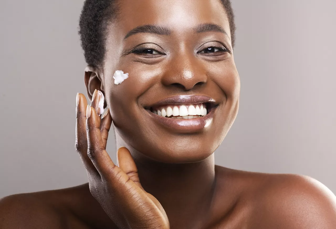 Skin Care Concept. Closeup Portrait Of Happy African American Woman Applying Moisturizing Cream on Face Over Grey Background