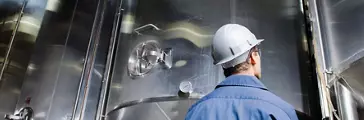 Man wearing white hard hat, safety glasses and blue uniform examines large steel vats