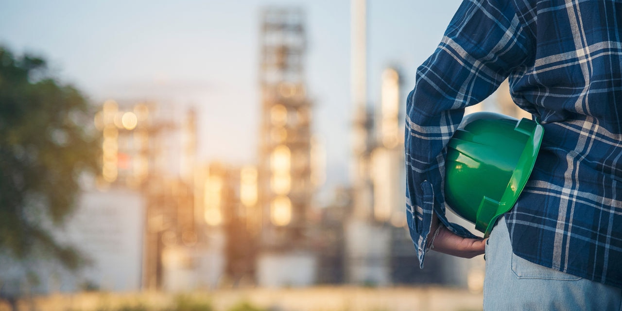Person holding green hard hat looks at manufacturing facility in the distance