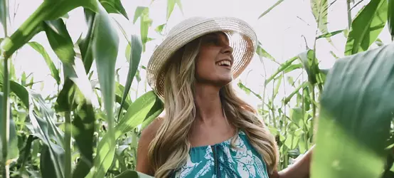 Young blonde woman wearing dress and hat walking through a corn field.
